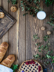 pie with berry filling, baguettes, glass of milk on a brown wooden background with flowers, green leaves, wooden desk, walnuts, top view