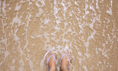 Fototapeta premium Top view of woman feet wearing a blue slipper on ocean sandy beach, relaxing concept
