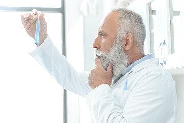 senior bearded scientist in white coat thinking and looking at tube with reagent in chemical lab