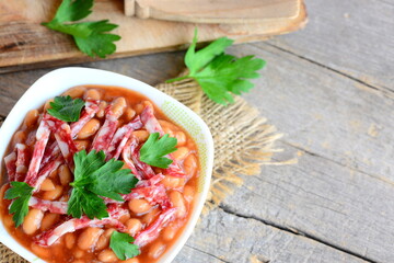 Smoked sausage and white bean stew. Homemade stewed white beans with smoked sausage, tomato sauce and parsley in a bowl and on a wooden background with copy space for text. Slow cooker white bean
