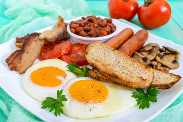 Traditional English breakfast: bacon, mushrooms, eggs, tomatoes, sausages, beans, toast on a white plate on a bright wooden background.  England classic cuisine.