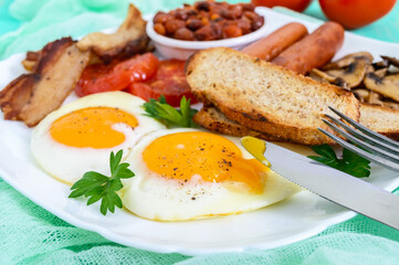 Traditional English breakfast: bacon, mushrooms, eggs, tomatoes, sausages, beans, toast on a white plate on a bright wooden background.  England classic cuisine. Close up