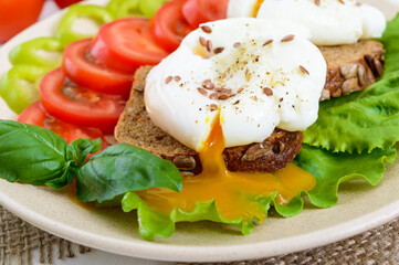 Sandwich with egg poached, lettuce, black bread with seeds, tomatoes, sweet pepper on a plate on a white wooden table. Close up