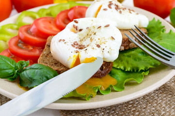 Sandwich with egg poached, lettuce, black bread with seeds, tomatoes, sweet pepper on a plate on a white wooden table. Close up