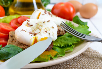 Sandwich with egg poached, lettuce, black bread with seeds, tomatoes, sweet pepper on a plate on a white wooden table. Close up