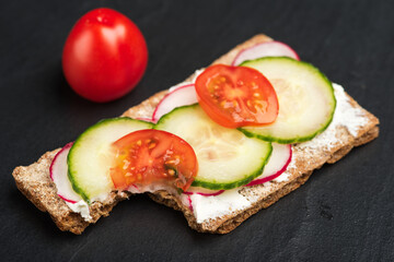 Crisp bread with vegetables. A bite is taken from the slice. Radish, cucumber and tomatoes on top of spread cheese garnish the bread. Tomato in background.