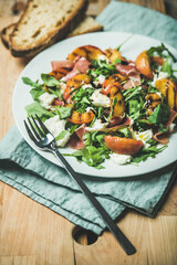 Arugula, prosciutto, mozzarella and grilled peach salad in white plate over blue napkin and wooden board, selective focus