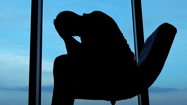 Silhouette of sad, unhappy businessman sitting on armchair by the window, against the sky
