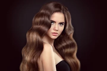 Wall murals Hairdressers Healthy hair. Brunette girl portrait with long shiny wavy hair. Beautiful model with curly hairstyle and eyeliner makeup isolated on studio black background.