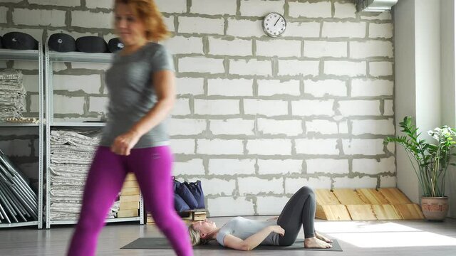 Middle-aged woman does yoga in a bright space with a brick wall and does stretching. Woman of 30-35 years of age does gymnastics. A female instructor helps a woman to perform exercises.