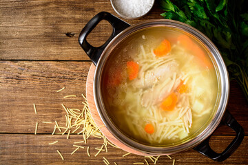 Homemade chicken soup with noodles and vegetables in metal pan on wooden table.