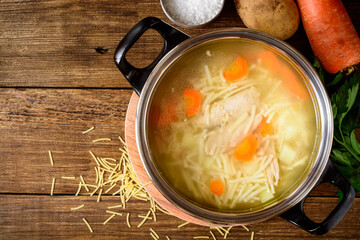 Homemade chicken soup with noodles and vegetables in metal pan on wooden table.