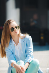 Beautiful young,stylish woman with,long straight blond hair,sitting on the sidewalk near his office in the open air in summer,dressed in a white shirt and pant suit blue in color,wearing sun glasses