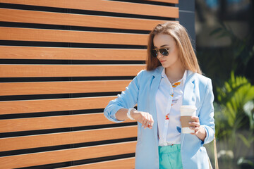 A stylish,young, beautiful woman in a blue trouser suit,white shirt,with a beige handbag,light long straight hair, sun glasses,posing near office outdoors in the summer with a paper cup of cappuccino