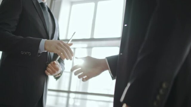 PAN shot with mid-section of businessmen standing before panoramic window, shaking hands and discussing information on clipboard 