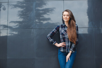 A young leader, a beautiful modern woman, a brunette with long straight hair and brown eyes,is dressed in a plaid shirt, posing standing outdoors standing near the mirror wall of the office