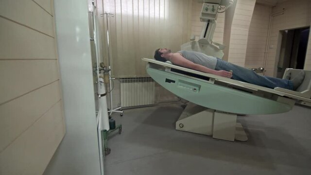 Middle aged male radiologist using scanner to performs x-ray imaging on younger man who lying on CT computed tomography machine at office, room interior, concept medical diagnostic, steady cam shot.