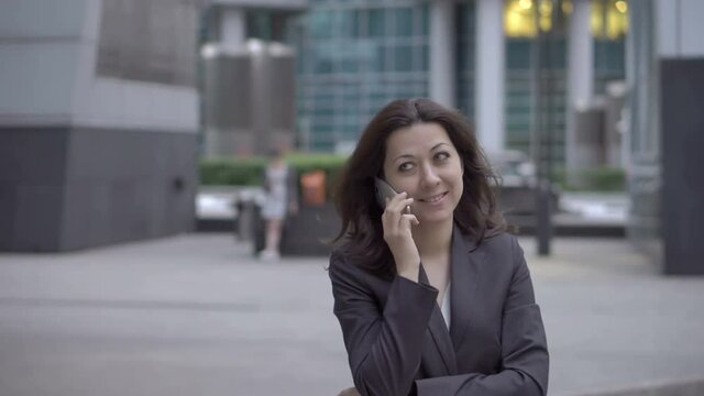 Attractive woman in business clothes approaching to the camera while talking on a mobile phone on the background buildings business centers, UHD 4K