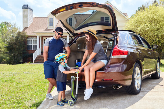 Family sits in cars trunk near house. Mother, father, son.
