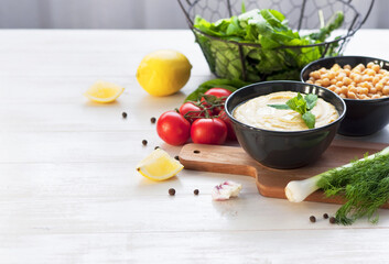 Bowl with hummus, chickpeas, lemon, cherry tomatoes and herbs on a white rustic wooden background. Middle Eastern cuisine. Background hummus with place for text.