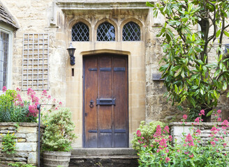 Old dark brown wooden doors in limestone cottage surrounded by flowers and plants .
