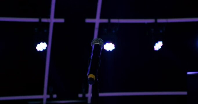Bluish violet lights shine on to a microphone. There is no performer present and as the lights shift the stage and equipment are illuminated by their radiant colors.
