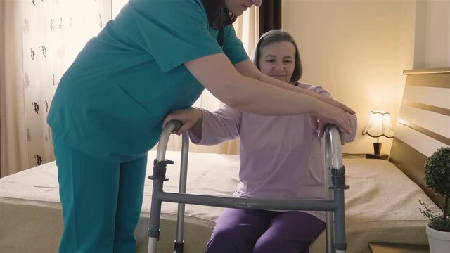 Friendly caregiver helping senior woman getting up from bed and walk with a walker. Home or hospice nursing and assistance concept. 4K footage at 60fps.
