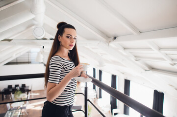 Close up portrait of a girl with brown hair holding take away coffee cup and smartphone while looking at the camera.