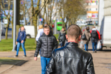 A young man with a short hairstyle and colored earrings in a black leather jacket walks away. View from the back