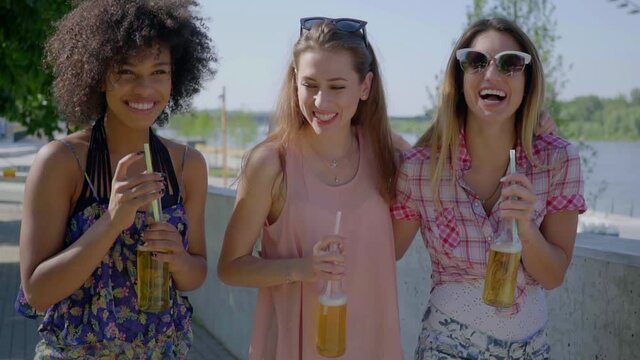 Three black and white women walking in city and laughing while having beer with straws.