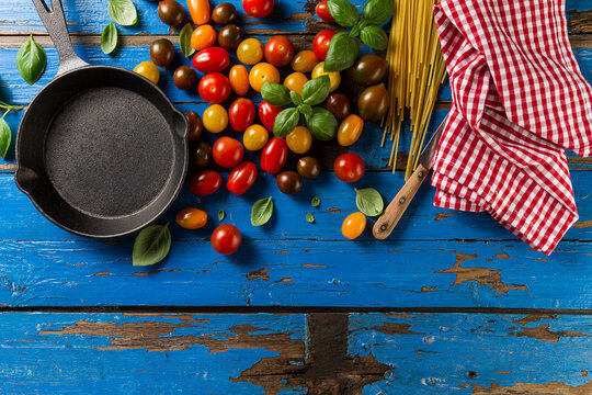Tasty fresh appetizing variety of tomatoes, basil, spaghetti, kitchen towel and pan on vibrant blue wooden background. Top View. Cooking process. Italian cuisine.
