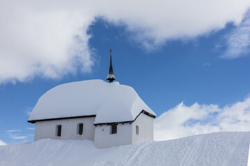 Fototapeta na wymiar Blue sky and clouds frame the alpine church immersed in snow Bettmeralp district of Raron canton of Valais Switzerland Europe
