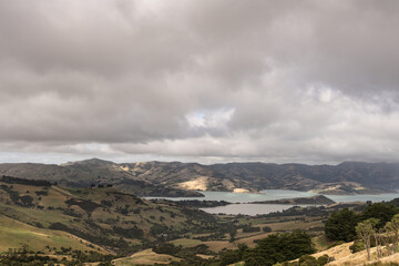 Fototapeta na wymiar Akaroa, New Zealand - March 14, 2017: The Akaroa Sound is an inlet from Pacific ocean in the remnants of a Miocene volcano. Heavy cloudscape and from the higher up hills. Vegetation in several shades.