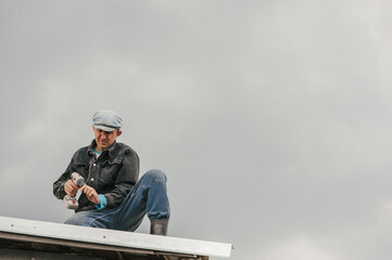 A man in work clothes tighten screws with a screwdriver on the roof against a cloudy sky.