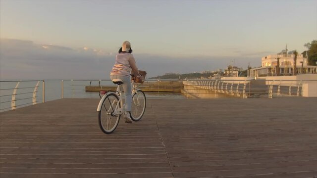 beautiful young woman wearing pink bomber, cap and sneakers riding retro white bicycle near sea or ocean on sunrise background