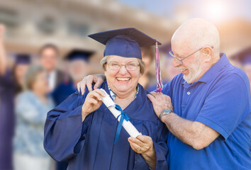 Senior Adult Woman In Cap and Gown Being Congratulated By Husband At Outdoor Graduation Ceremony.