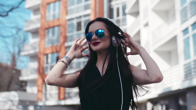 Young hipster girl in sunglasses listens to music on headphones, and happily dances to the rhythm from side to side in the sunny street. Fashionable outfit, red lips, shorts. Joyful mood.