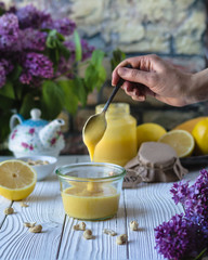lemon curd in a glass jar with woman's hand holding a spoon, lemons, nuts, tea pot  and lilac