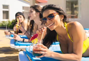 Three Beautiful girls in swimwear and sun glasses are drinking cocktails through a straw while sunbathing on the chaise longue near the pool. One girl is smiling and looking at camera