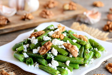 Vinegar and oil green bean salad recipe. Delicious green string beans salad with cottage cheese, peeled walnuts, garlic and spices on a white plate and wooden table. Warm salad for healthy dinner idea