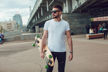 Stylish man in sunglasses and with a beard stands on the street with a long board. T-shirt mock up. © kanashkin