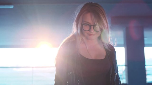 Gorgeous blonde long-haired young woman walking down the parking, turning to camera and seductively smiling in a bright sunshine. Modern nerdy style, hipster. Natural beauty, playful mood.