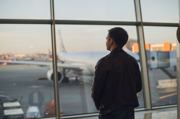 Fototapeta premium Young man is standing near window at the airport and watching plane before departure. Focus on his back