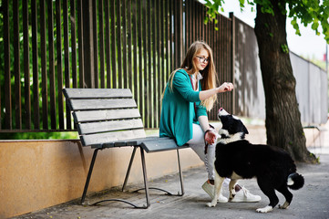 Naklejka na ściany i meble Trendy girl at glasses and ripped jeans with russo-european laika (husky) dog on a leash, against bench on street of city. Friend human with animal theme.