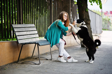 Naklejka na ściany i meble Trendy girl at glasses and ripped jeans with russo-european laika (husky) dog on a leash, against bench on street of city. Friend human with animal theme.