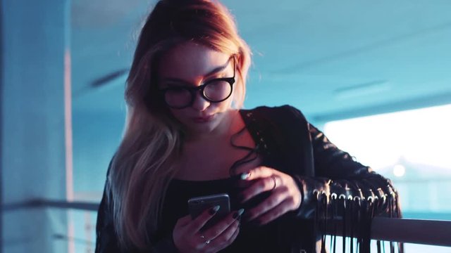 Attractive nerdy-like blonde woman in a black boho-style leather jacket using her phone for the Internet surfing, corrects eyeglasses, looks around. City on the background.