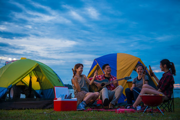 Camping of happy asian young travellers at lake, asian man and women group, relaxing, sing a song and cooking, at sunset.