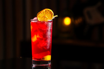 Closeup glass of cocktail with strawberry syrup, rum and lemon decorated with orange slice at bar background.