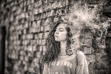 Beautiful vape teenager. Portrait of a pretty young white girl with curly hair vaping an electronic cigarette near old destroyed brick wall. Black and white. Toned. Lifestyle.