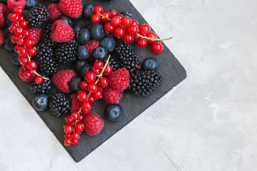 Mix of berries raspberries red currants blueberries and blackberries on black slate board. White stone background.  Overhead view and copy space